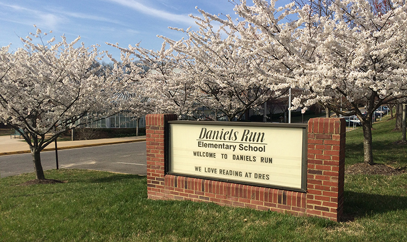 Photograph of the Daniels Run Elementary School sign that faces Old Lee Highway. The sign is mounted in brick housing and reads Welcome to Daniels Run, we love reading at DRES. The cherry trees on the school grounds are in full bloom with white blossoms, obscuring the view of the front of the school building.